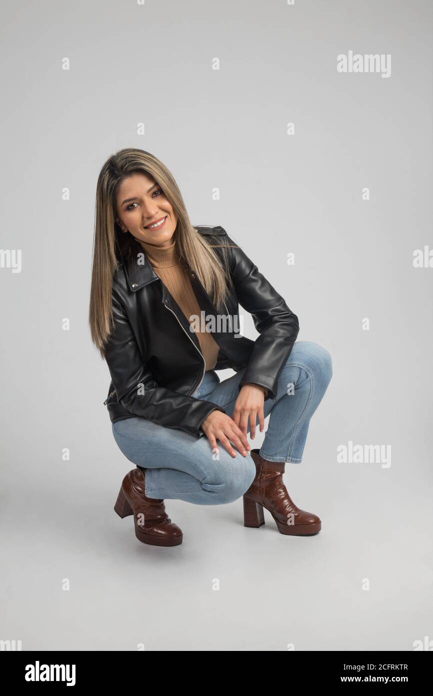 Latino femme s'accroupir avec une tenue décontractée qui ont une veste en cuir  noir, un Jean et des bottes sur un fond gris, studio Photo Stock - Alamy