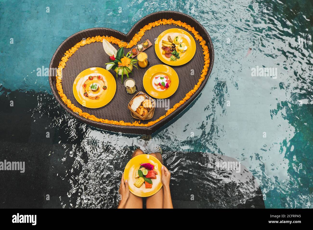 Petit déjeuner flottant dans la piscine. Femme mains tenant la plaque avec des fruits frais. Régime alimentaire sain d'été. Style de vie tropical. Plateau en rotin, fleurs orange, style Bali et service. Banque D'Images