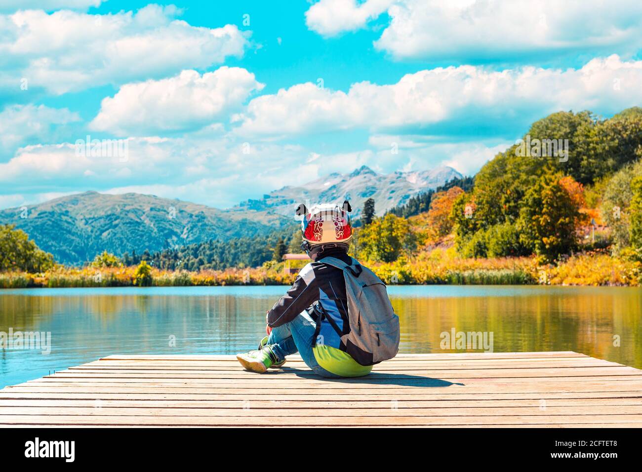 Paysage d'automne. Une personne dans une veste et un casque de moto, assise sur la jetée sur le fond du lac et de la forêt d'automne. Ciel bleu avec Banque D'Images