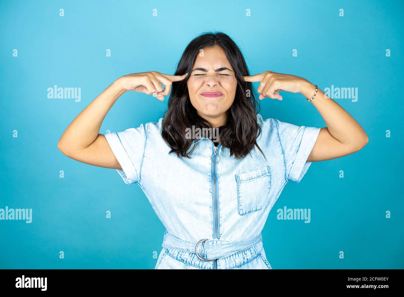 Jeune belle femme portant une combinaison en denim sur fond bleu isolé avec la main sur la tête pour la douleur dans la tête parce que le stress. Souffrir de migraine. Banque D'Images