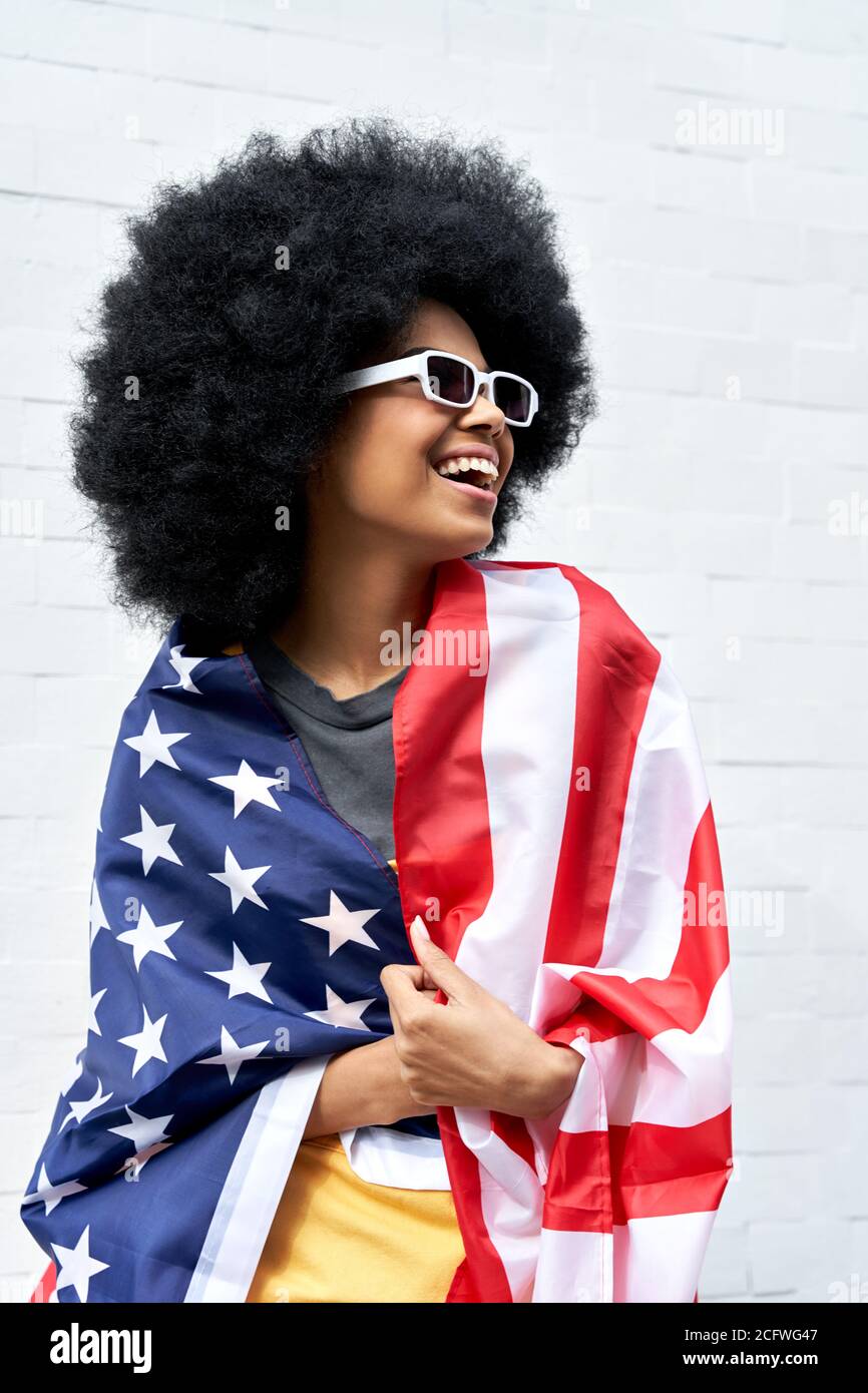 Bonne jeune femme africaine enveloppée dans le drapeau américain debout contre le mur blanc. Banque D'Images