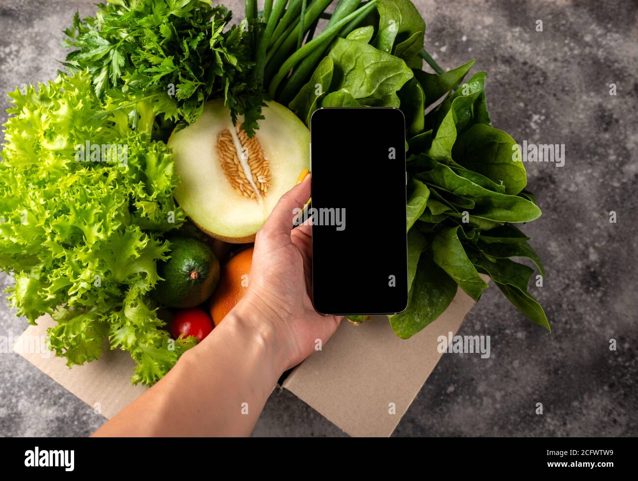 Smartphone dans la main féminine et boîte complète de légumes biologiques de la ferme. Commander livraison de nourriture en ligne concept. Banque D'Images