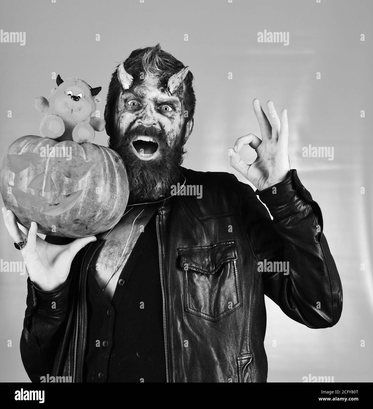 Le démon au visage choqué tient la Jack o lanterne sculptée et montre le signe ok. Monstre avec décorations d'octobre. Concept de fête d'Halloween. L'homme portant le maquillage tient le jouet de citrouille et de diable rouge sur fond gris Banque D'Images