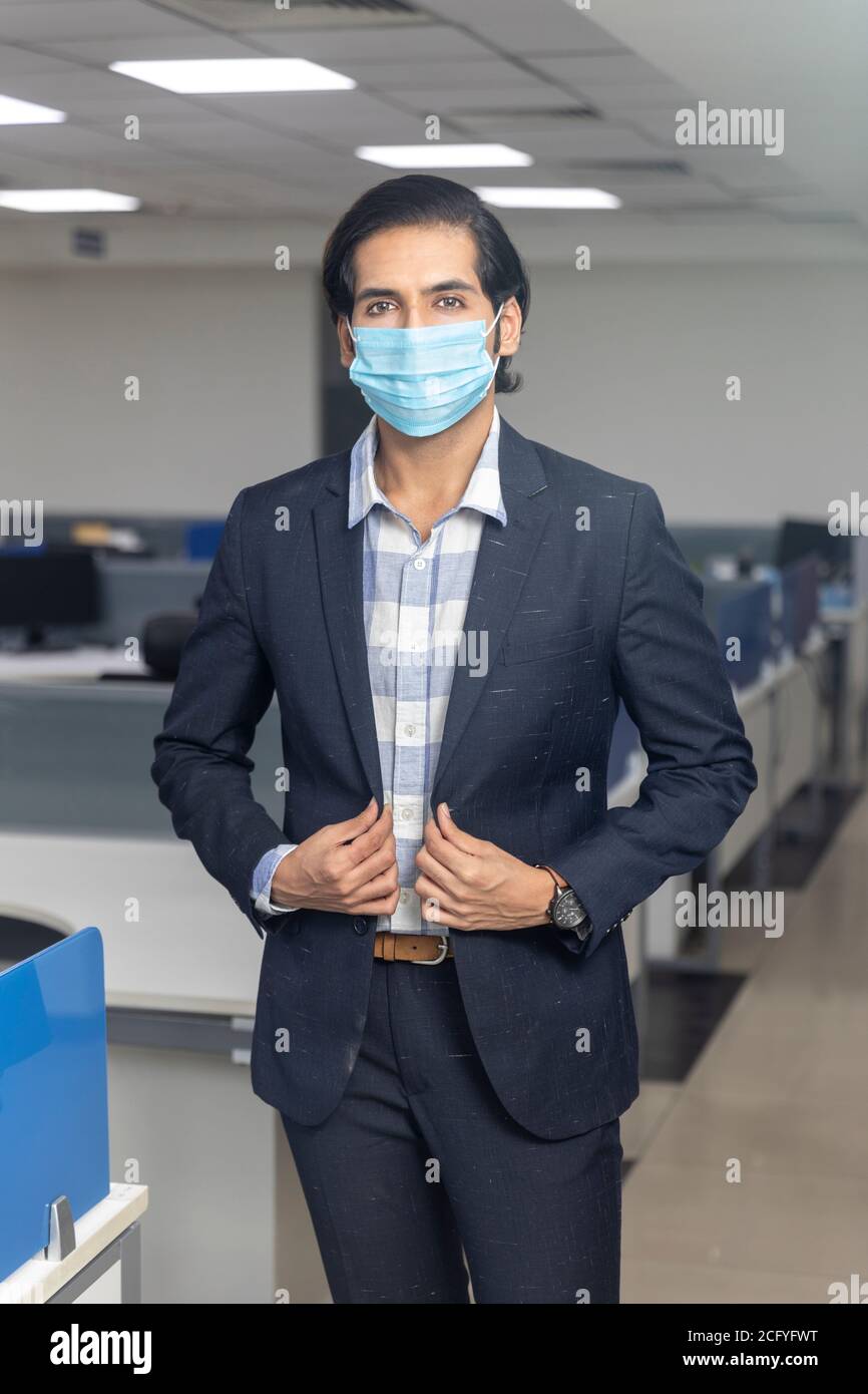 Portrait d'un jeune homme d'affaires indien élégant portant un masque de protection Covid, debout contre le fond du bureau. Environnement de bureau d'entreprise. Banque D'Images