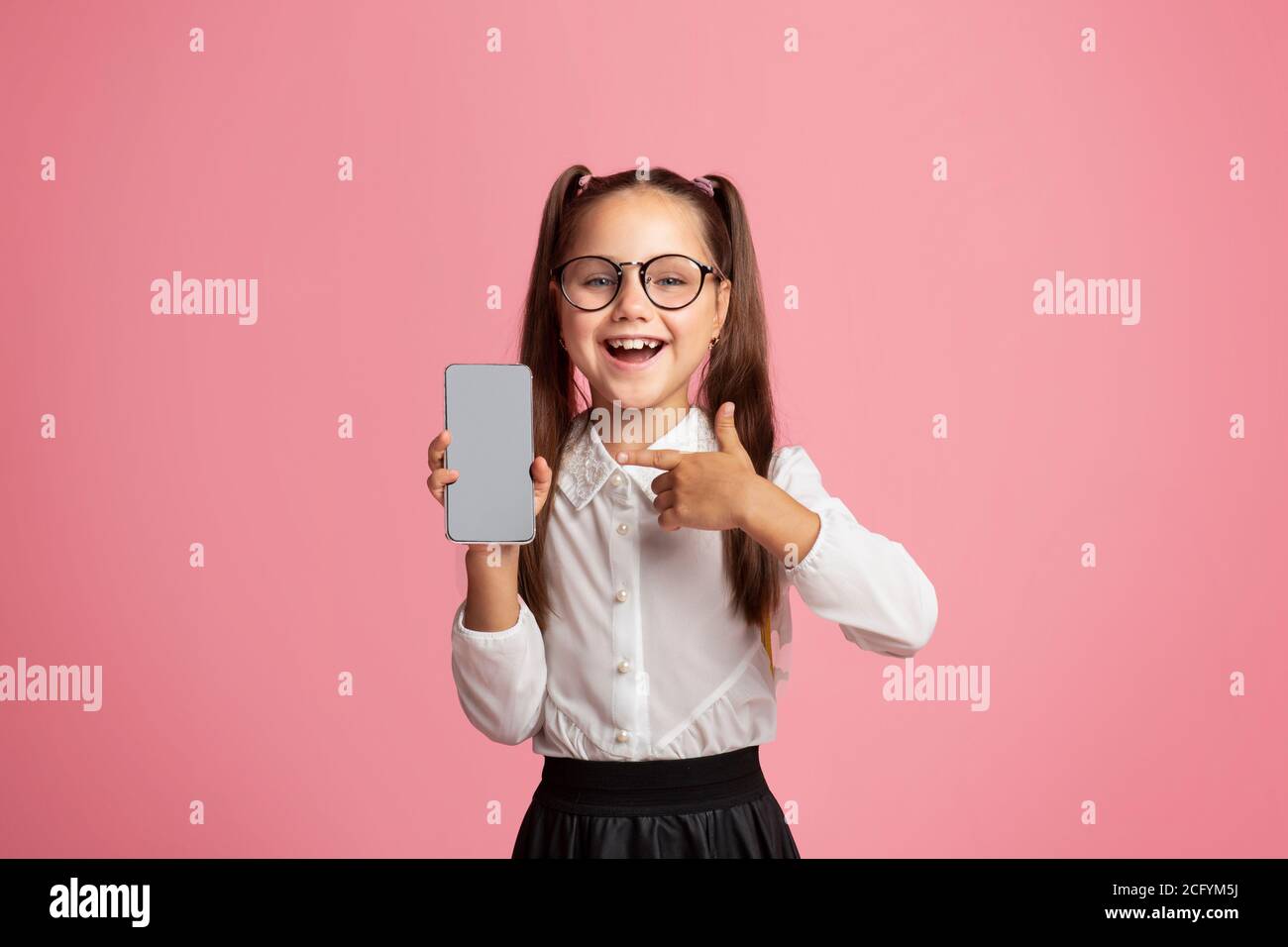 Bonne écolière dans des verres à bouche ouverte montre le pouce vers le haut et smartphone avec écran vide Banque D'Images