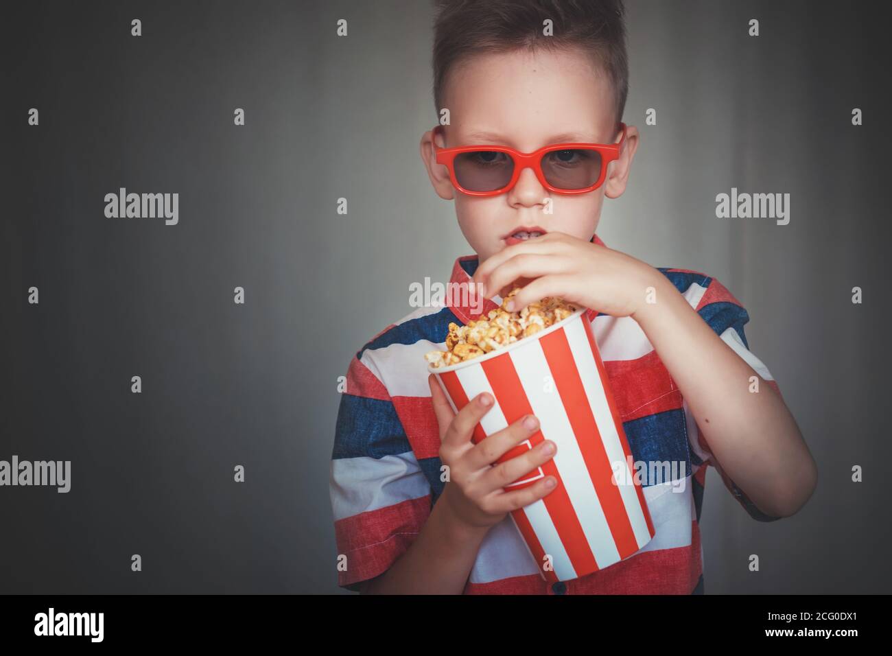 Un jeune garçon regarde un film en lunettes 3D au cinéma ou à la maison. Un  petit enfant mange du pop-corn sur fond gris. Home Cinéma. Mignon enfant  dans un cinem vintage