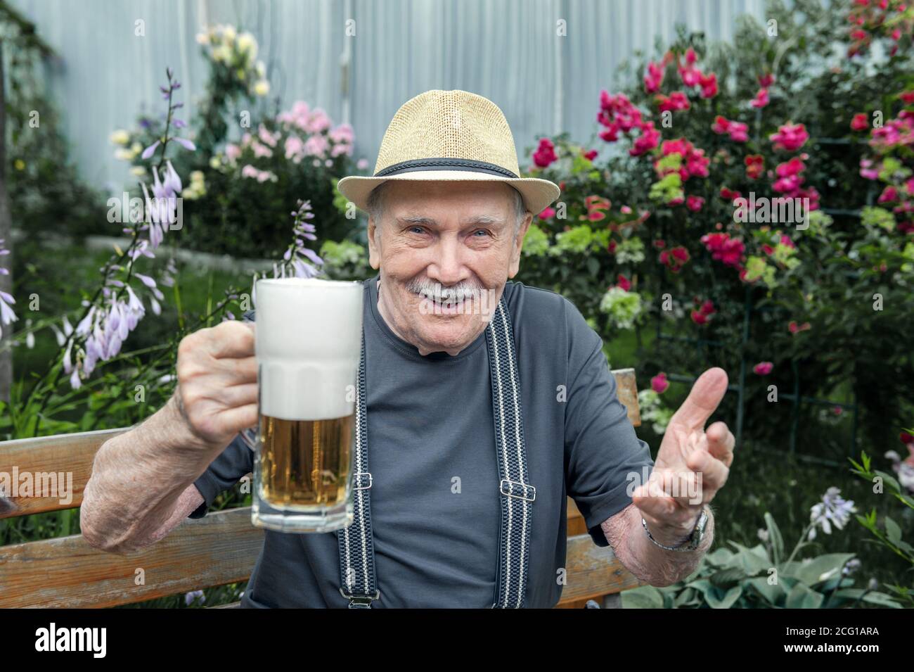 un homme âgé au chapeau boit de la bière à l'extérieur d'un pub sur un banc dans le jardin. Le vieil homme verse de la bière dans un verre. Banque D'Images