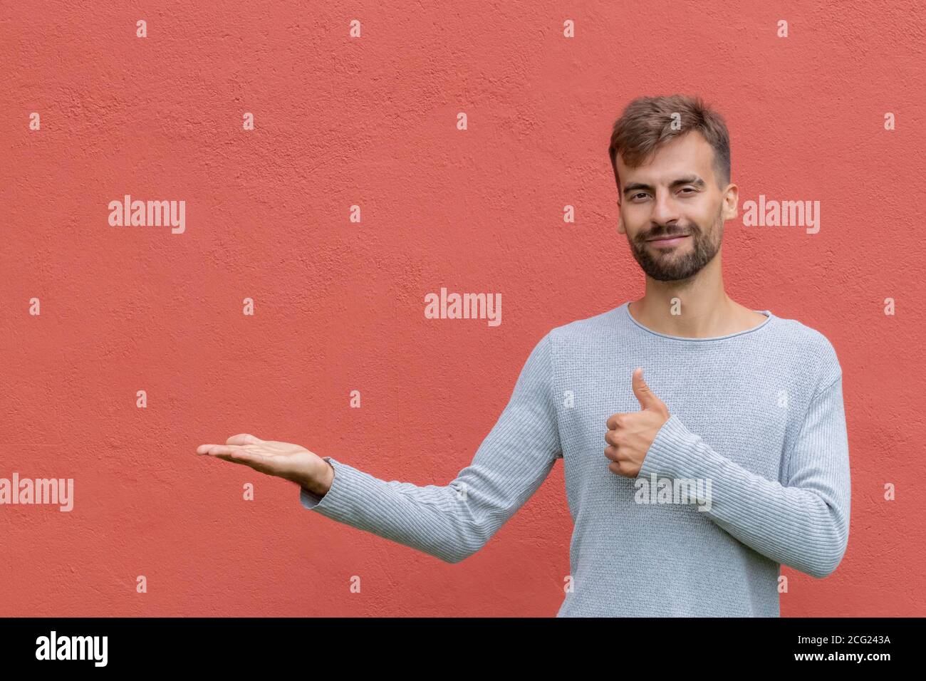 Jeune homme barbu souriant avec chemise grise tenant un espace de copie imaginaire sur la paume pour insérer une annonce sur un arrière-plan isolé de mur rouge. Copier la flèche Banque D'Images