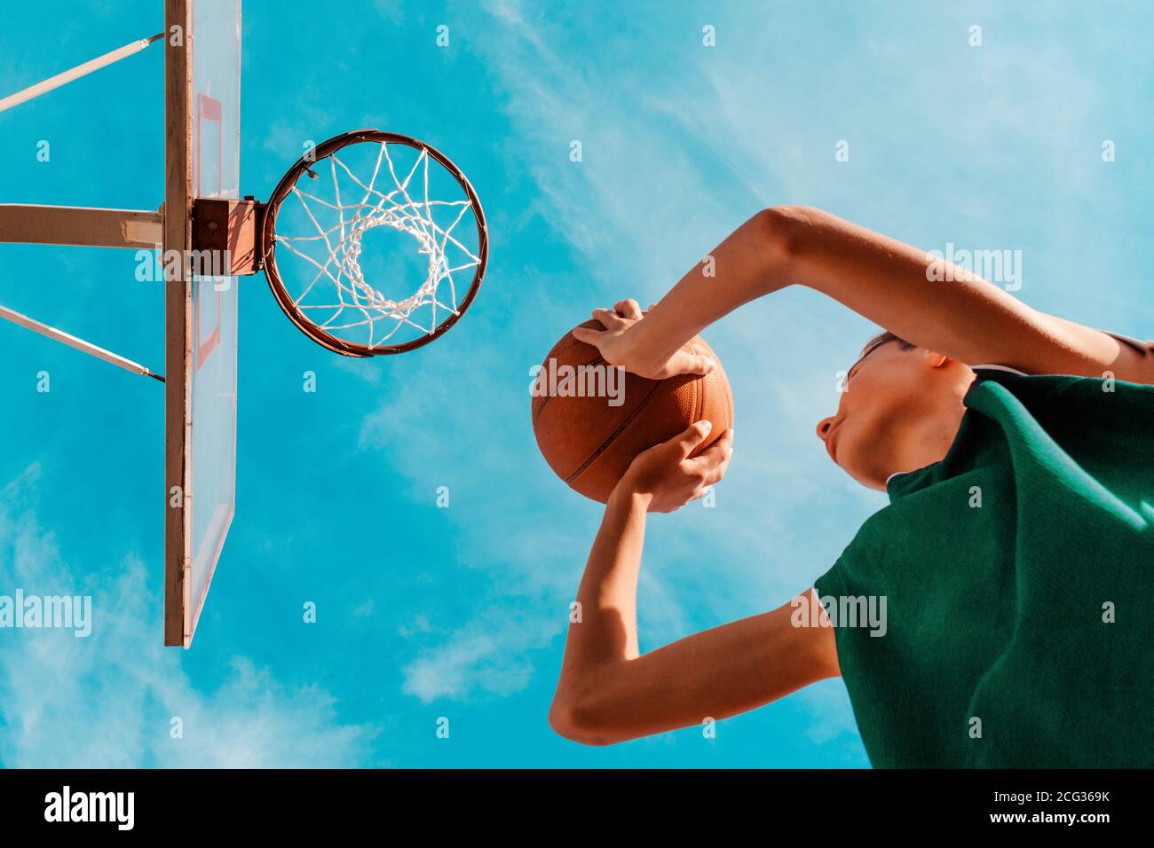 Sports et basket-ball. Un jeune adolescent en combinaison verte jette une balle dans le panier. Vue de dessous. Ciel bleu en arrière-plan. Banque D'Images