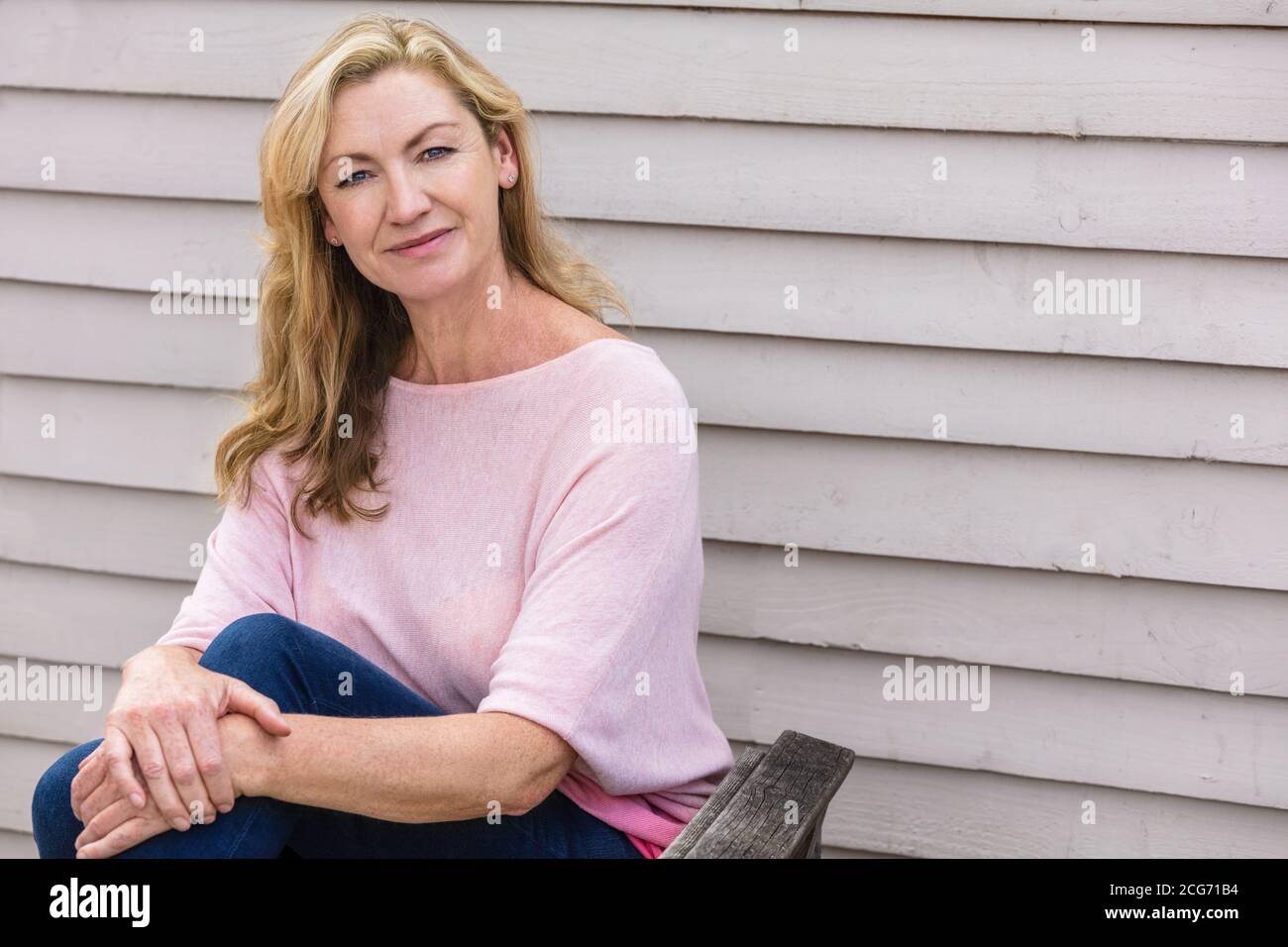 Portrait extérieur d'une jolie femme blonde d'âge moyen souriante assis sur une chaise de jardin en bois Banque D'Images