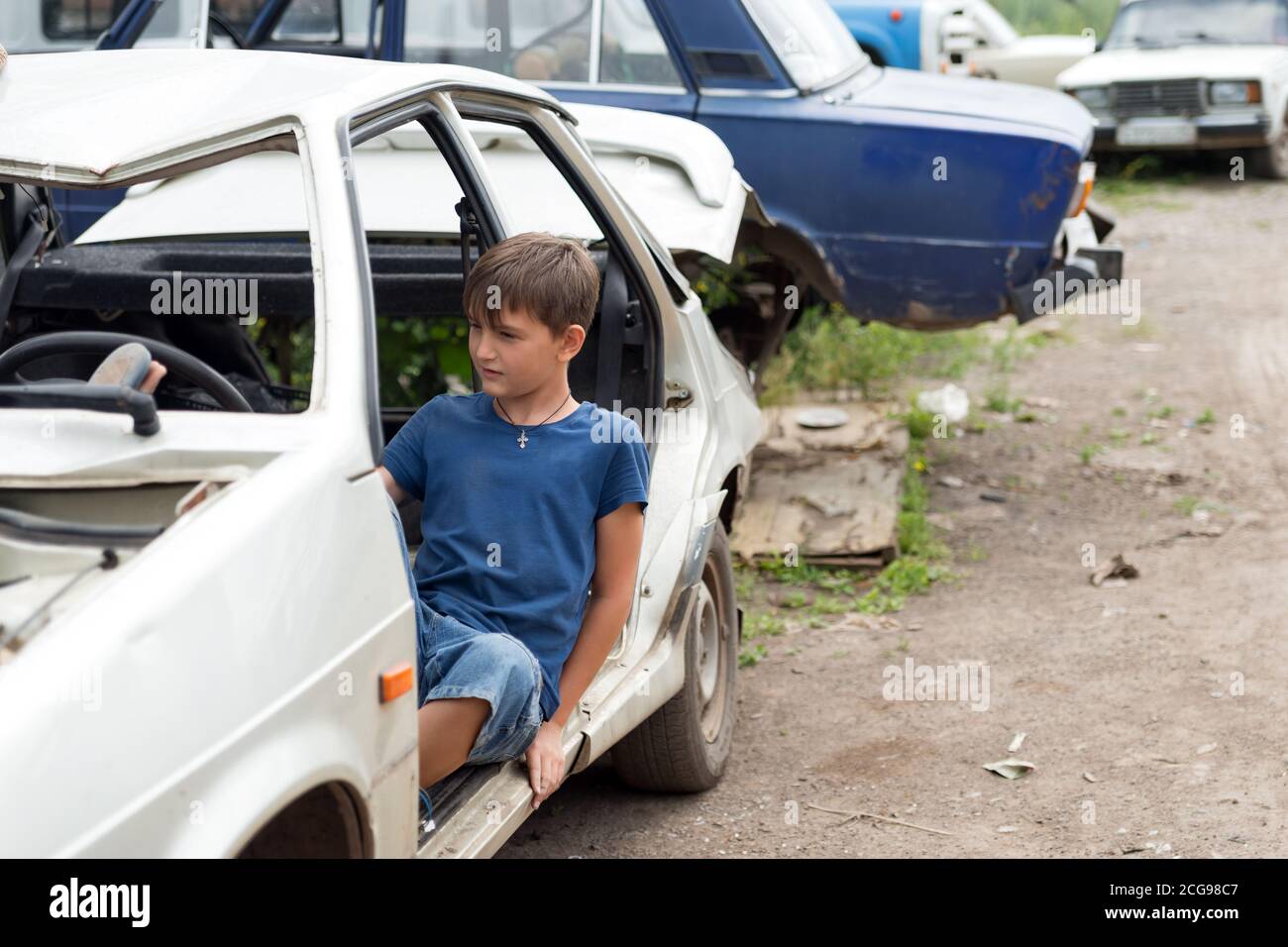 Un garçon de 11 ans est assis dans une voiture démontée dans un dépotoir de vieilles voitures abandonnées par une belle journée d'été. Banque D'Images