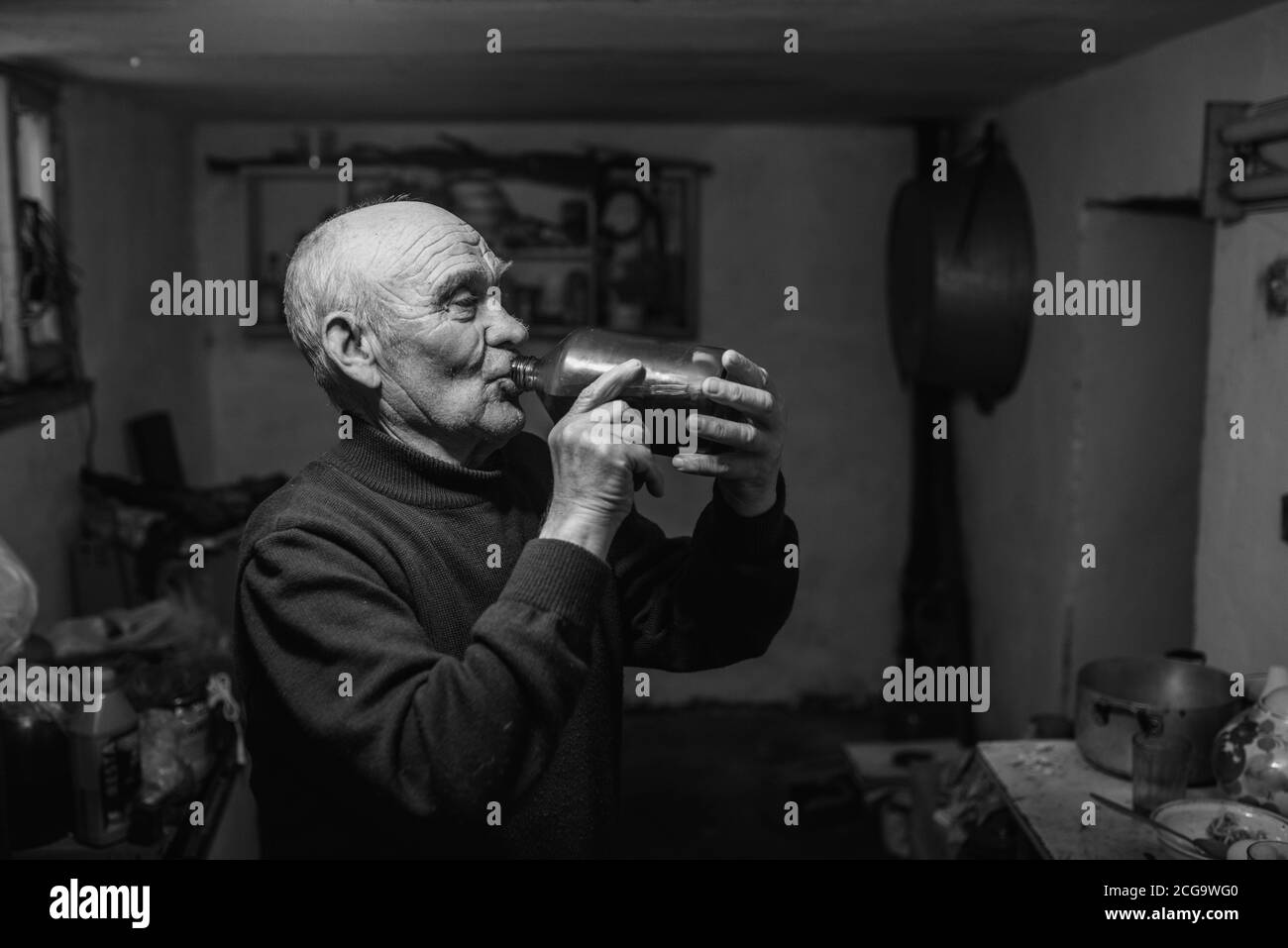 un vieil homme aux cheveux gris, aux rides profondes, boit du vin rouge fait maison dans une bouteille de verre au sous-sol sombre de la maison. Banque D'Images