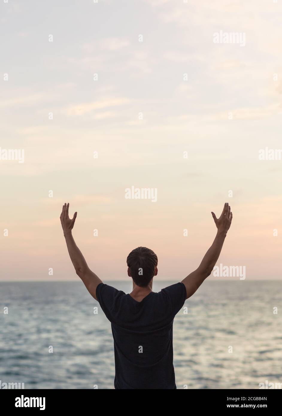 Jeune homme en bonne santé avec les bras vers le ciel célébrant pendant un beau coucher de soleil face à l'océan. Bien-être et liberté. Banque D'Images