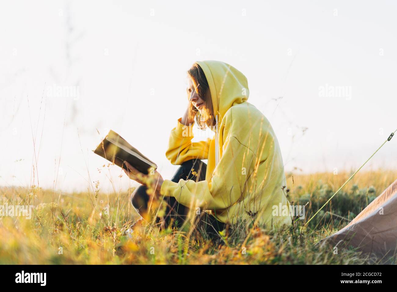 Jeune femme voyageur à capuchon jaune lisant le livre contre la tente. Tourisme local, week-end Banque D'Images
