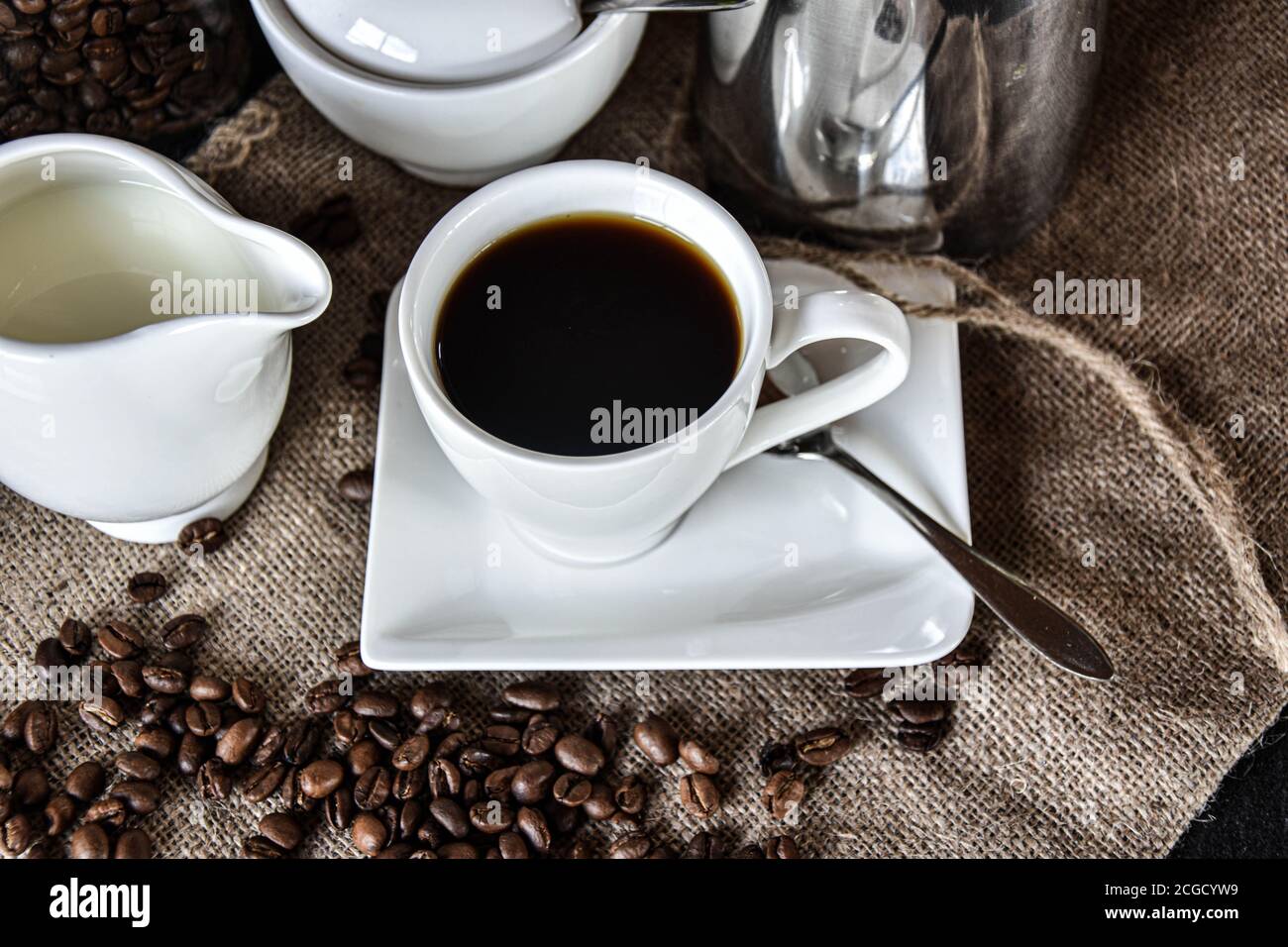 Une tasse de café avec de la vaisselle blanche comme un pot à lait, un pot à sucre et une soucoupe servis chauds sur une texture jute avec une décoration de grains de café Banque D'Images