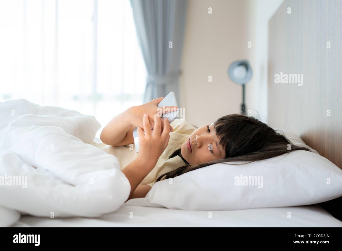 Bonne fille asiatique regardant le téléphone portable tout en étant allongé sur le lit dans la chambre à la maison le matin après le réveil se préparant à aller à l'école. Banque D'Images