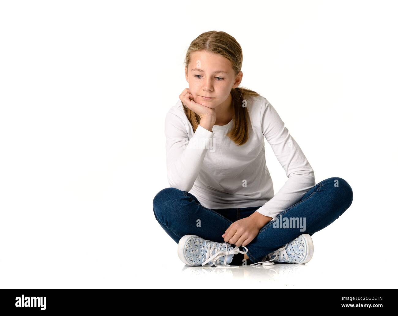 Jeune fille blanche blonde en chandail blanc, jenas et baskets assis sur fond blanc sans couture en studio Banque D'Images