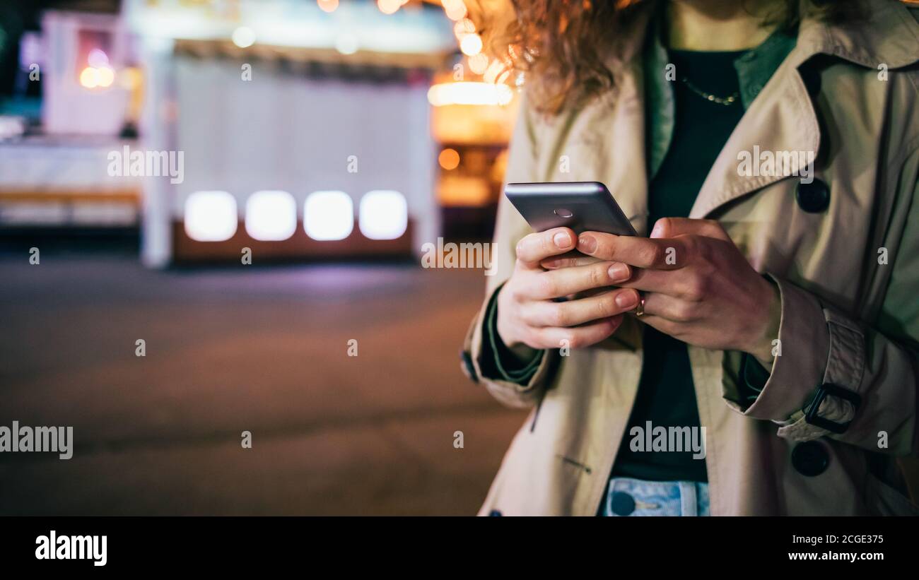 Gros plan jeune femme élégante utilisant un téléphone portable debout sur une rue de ville lumineuse de nuit, espace de copie. Banque D'Images