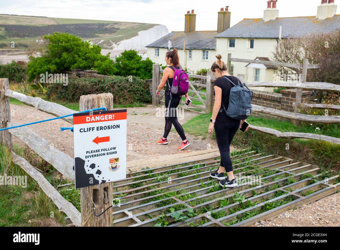 Les jeunes femmes marchent au-dessus de la grille de bétail à danger Cliff érosion panneau aux célèbres cottages de garde-côtes sur les falaises surplombant Cuckmere Haven Banque D'Images