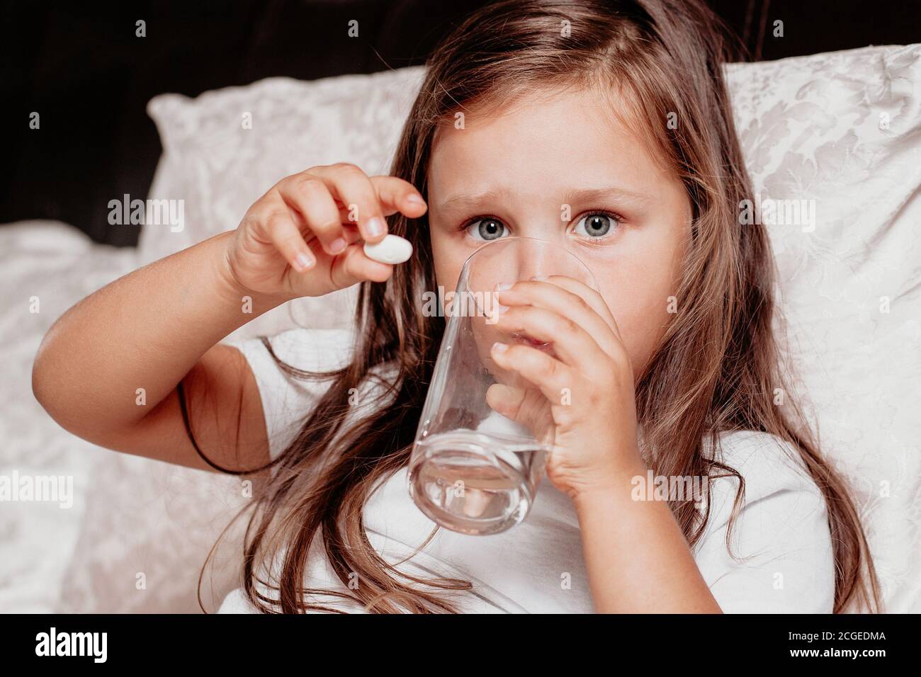 un enfant malade, une fille assise sur un lit avec un coussin blanc, en train de boire des médicaments contre le rhume Banque D'Images