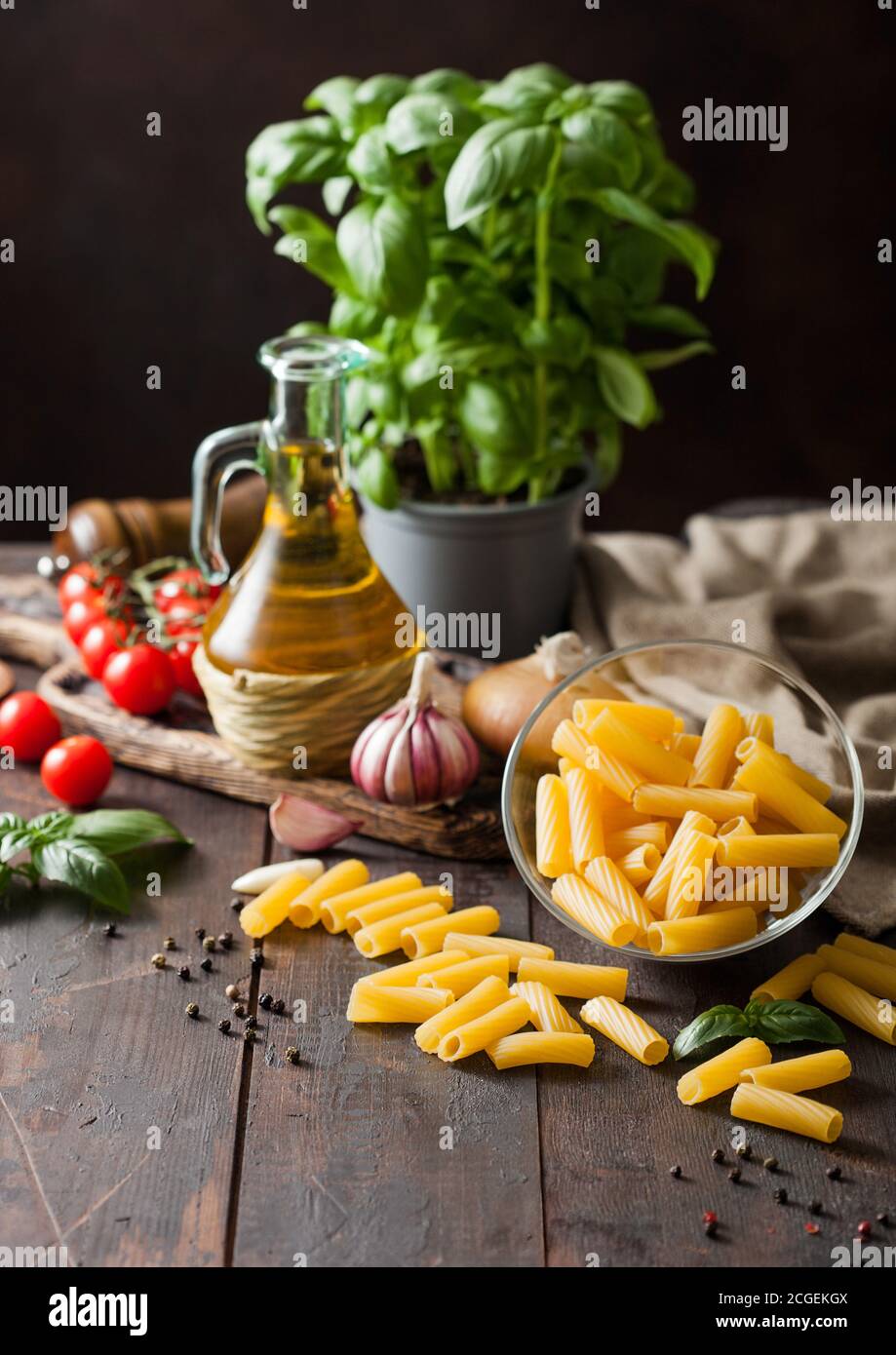 Pâtes de penne crues dans un bol en verre avec de l'huile et de l'ail, des plantes de basilic et des tomates, du poivre et du linge de maison sur fond de table en bois. Banque D'Images