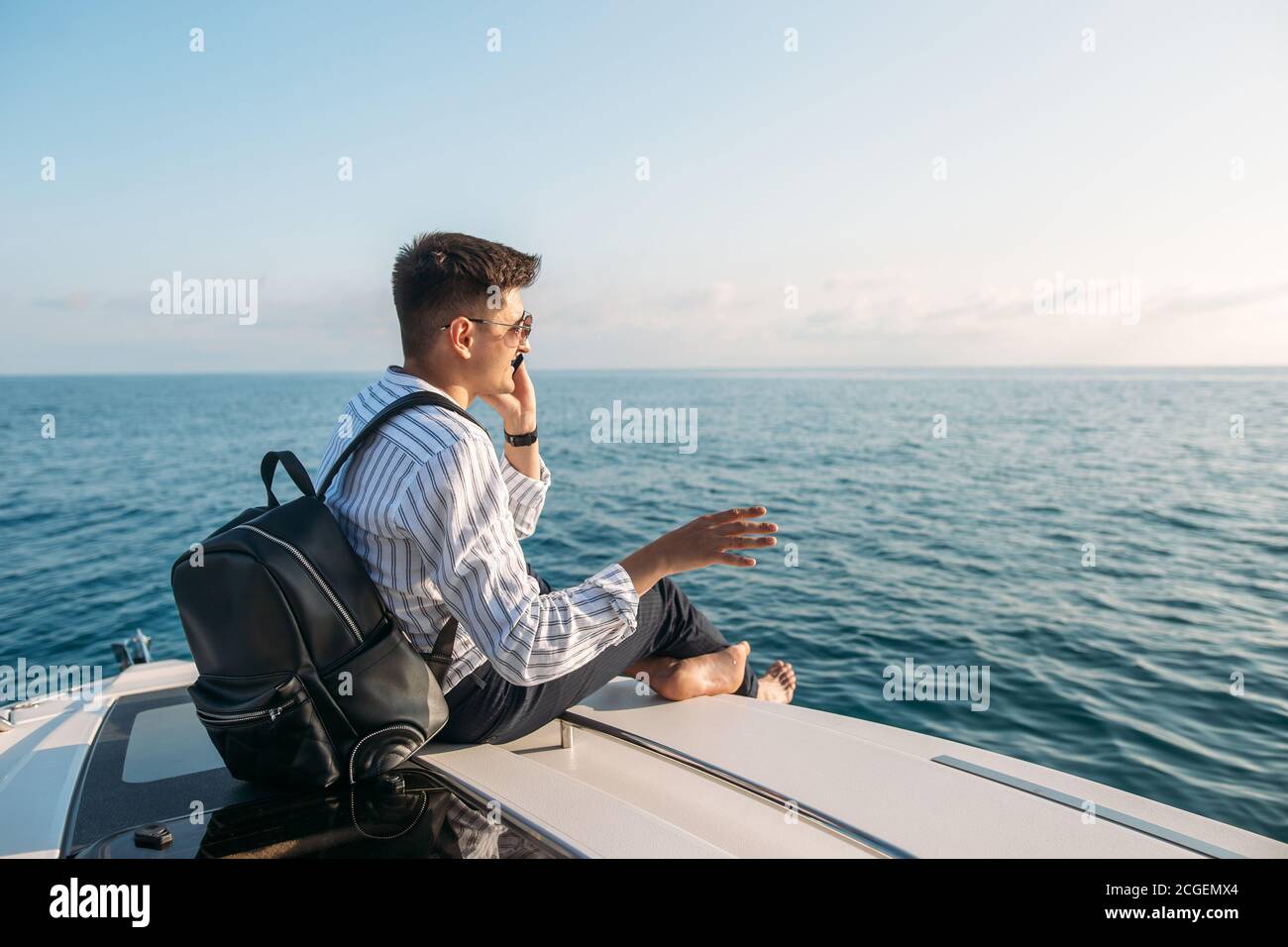 Espace pour le texte. Beau jeune homme avec sac à dos dans les mains, vêtu de vêtements de bureau relaxant et travaillant sur une tablette numérique sur un arc de bateau à moteur, au-dessus de la mer Banque D'Images