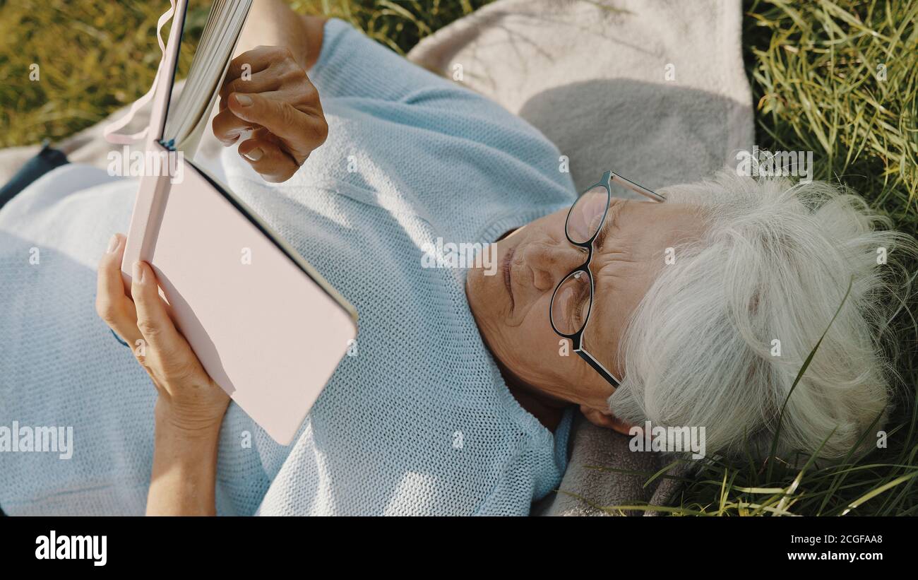 femme sénior aux cheveux gris lisant un livre tout en se reposant sur l'herbe dans la nature. Photo de haute qualité Banque D'Images