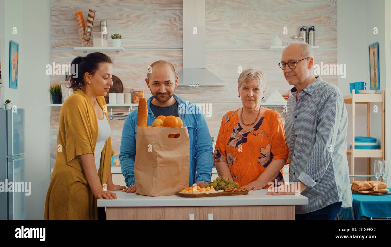 Portrait de famille au ralenti dans la cuisine moderne. Des gens heureux souriant à l'appareil photo dans la salle à manger autour du sac de papier avec des provisions regardant l'appareil photo Banque D'Images