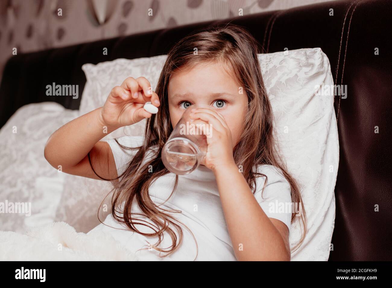 un enfant malade, une fille assise sur un lit avec un coussin blanc, buvant de l'eau d'un verre et tenant une pilule blanche à la main Banque D'Images