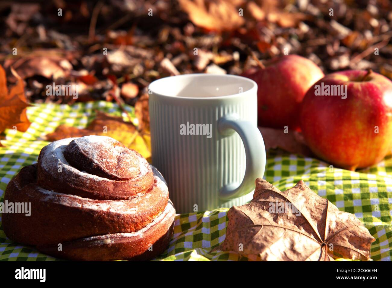 L'automne encore la vie. Pommes mûres rouges, une tasse de café avec du lait dans une tasse bleue, un pain à la cannelle doux sur une nappe à carreaux verts sur fond Banque D'Images