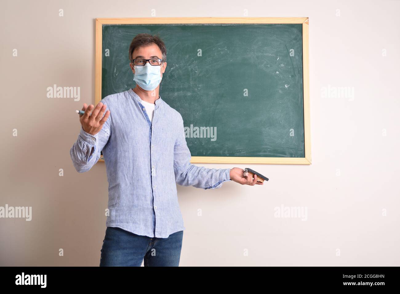 Professeur d'école primaire protégé avec masque en raison du risque de infection à coronavirus expliquant la leçon en classe devant a tableau noir à la craie verte Banque D'Images