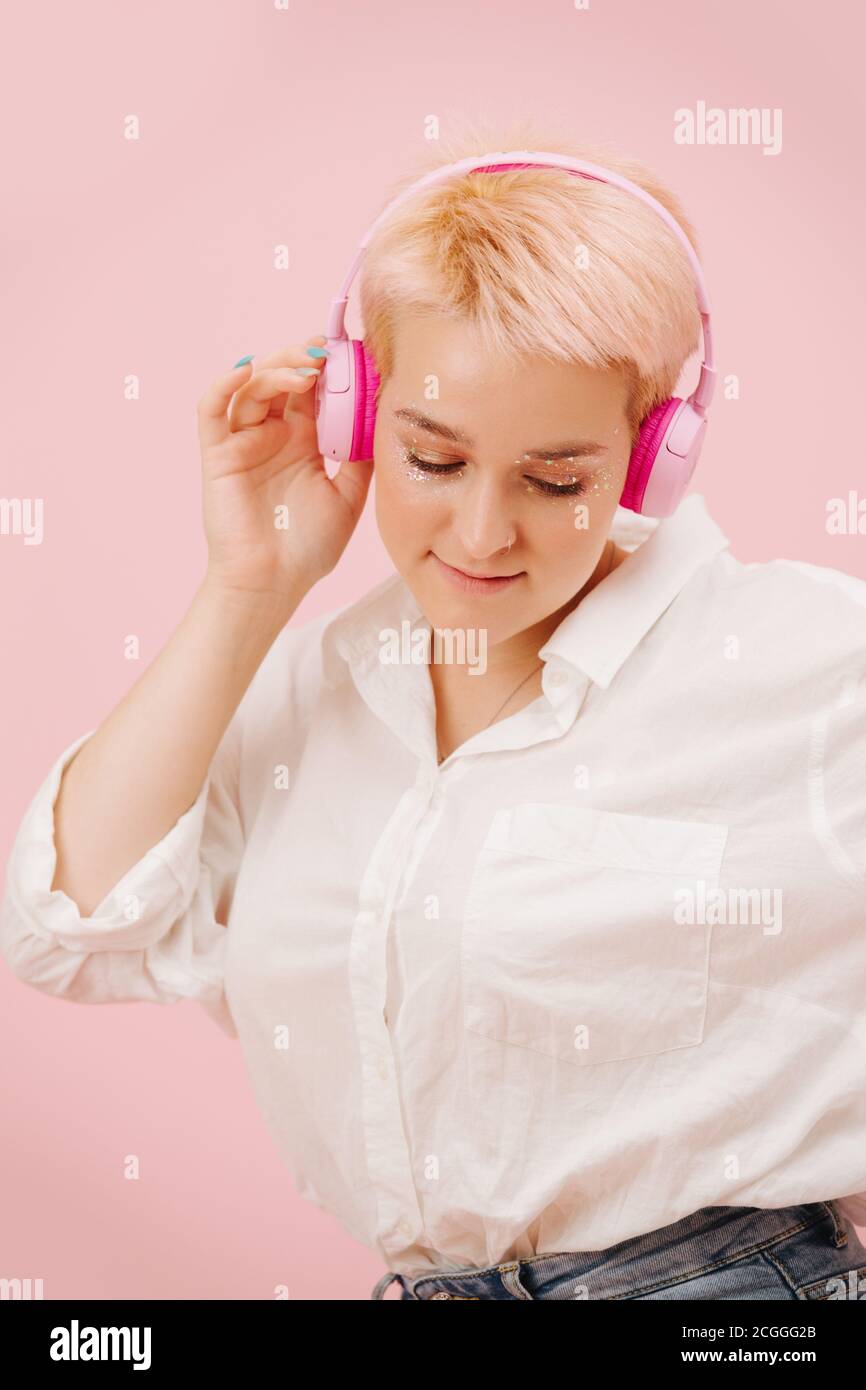 Jeune femme timide avec cheveux courts écoutant des écouteurs sans fil. Sur rose. Banque D'Images