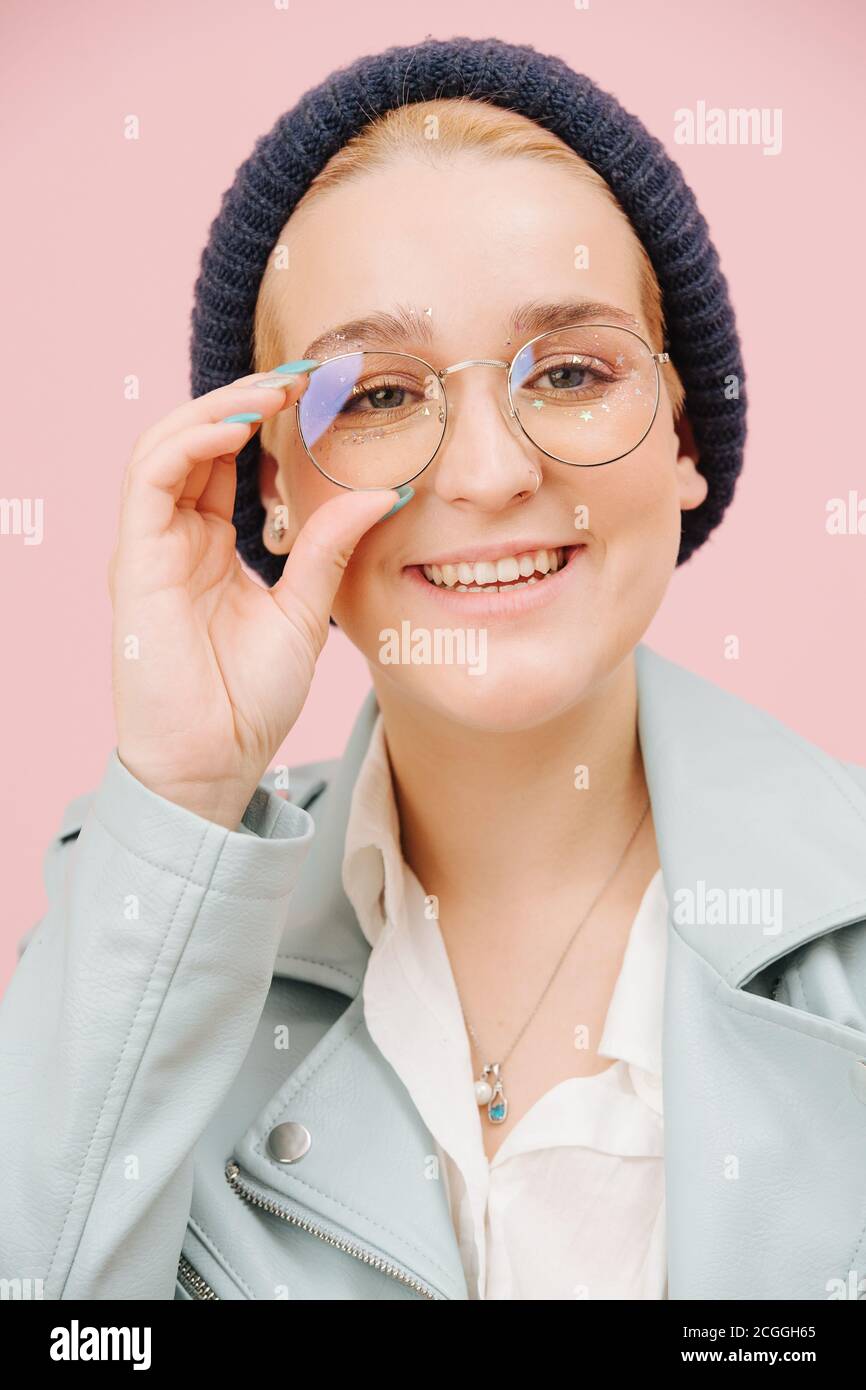 Jeune femme excentrique souriante dans un chapeau et des verres ronds souriant. Sur rose. Banque D'Images
