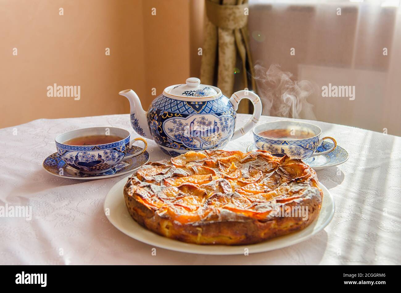 Tarte aux pommes sur un plat blanc, une théière et deux tasses de thé chaud sur une table recouverte d'une nappe blanche. Banque D'Images