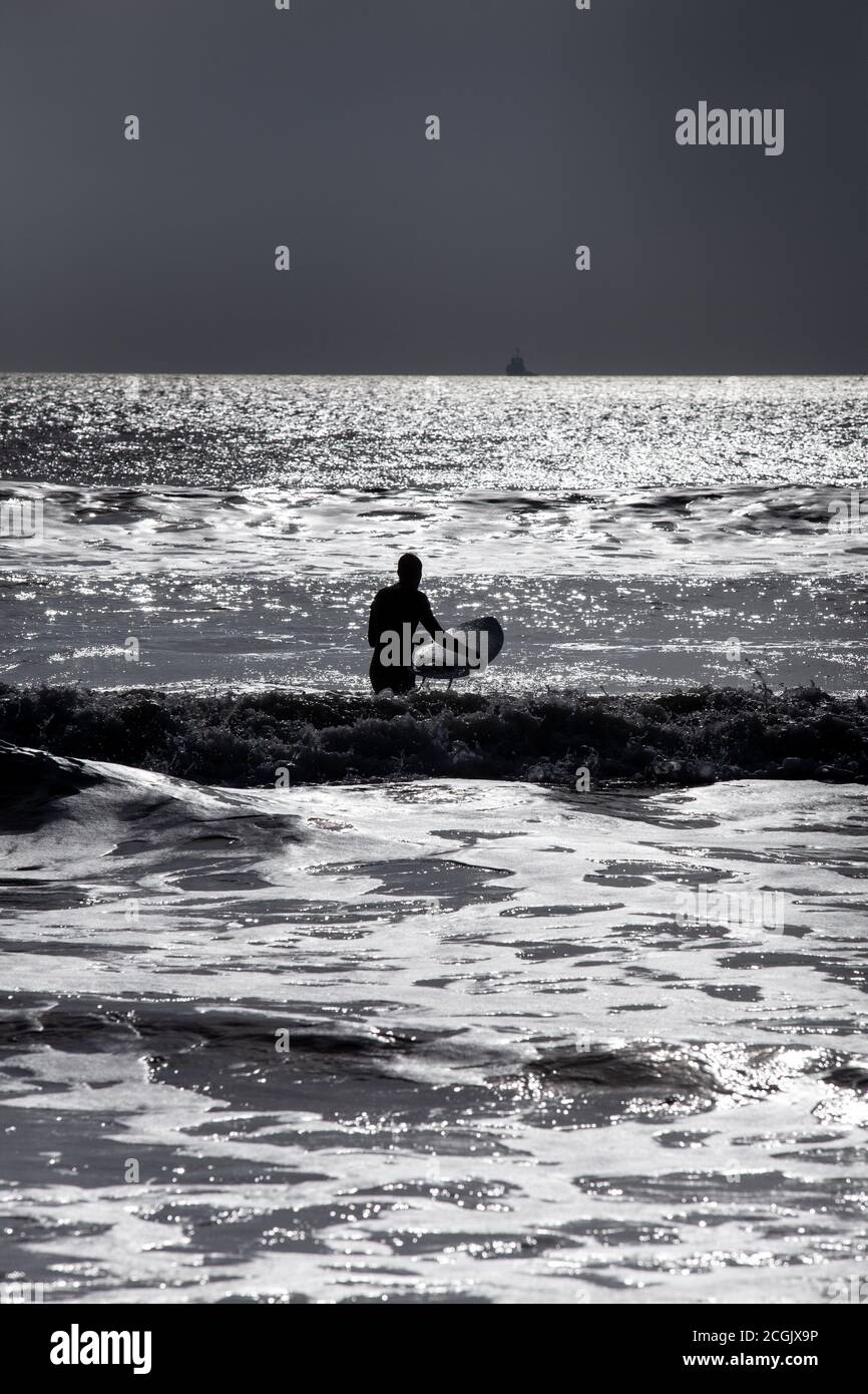 Le surfeur silhouetté se promène dans les petites vagues à côté de la jetée de la plage de Bournemouth pour profiter du soleil d'hiver et surfer avant le début de la pluie. Banque D'Images