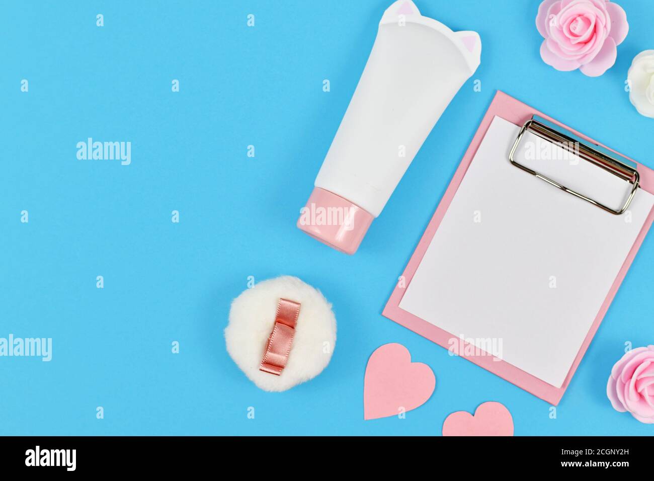 Plat avec bloc-notes vierge et fleurs roses, tube de crème en forme de chat, coeur de papier et poudreuse sur fond bleu avec espace de copie vide Banque D'Images
