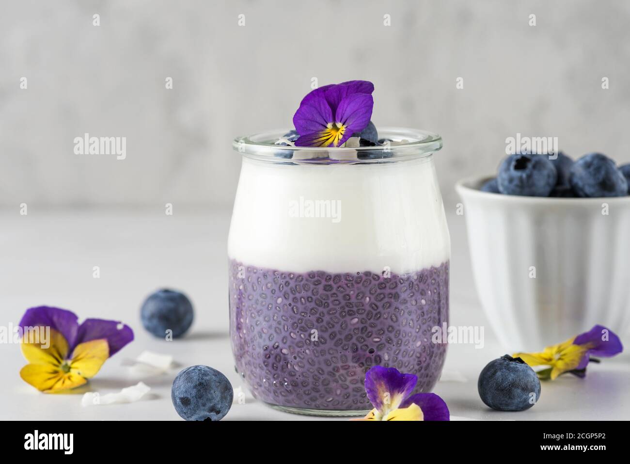 Petit déjeuner sain composé de parfaits aux myrtilles, de fruits frais, de yaourts à la noix de coco, de pudding au chia et de fleurs comestibles sur table blanche Banque D'Images