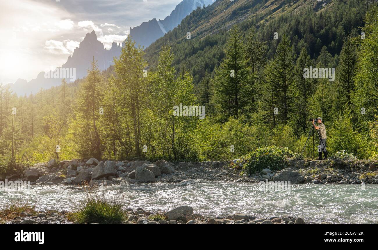 Photographe de la nature prendre des photos à l'aide d'un appareil photo professionnel à côté de Scenic Mountain River. Paysage alpin élevé en arrière-plan. Banque D'Images