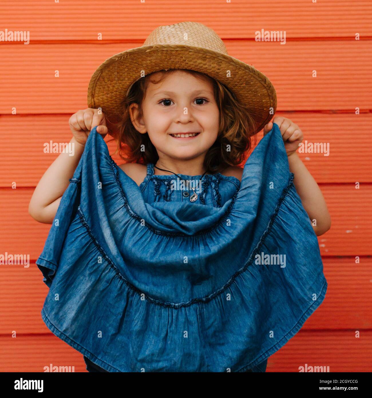 Bonne fille d'enfant en chapeau d'été et un beau bleu habillez-vous contre le mur rouge Banque D'Images