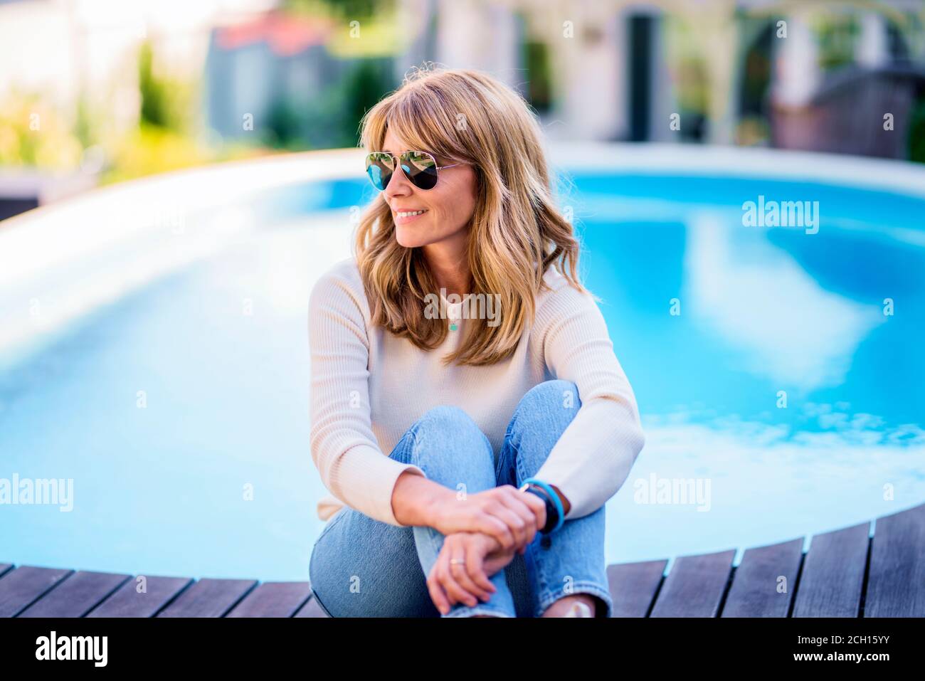Portrait d'une femme mûre heureuse portant des vêtements décontractés et des lunettes de soleil tout en se relaxant sur la piscine à la maison. Banque D'Images