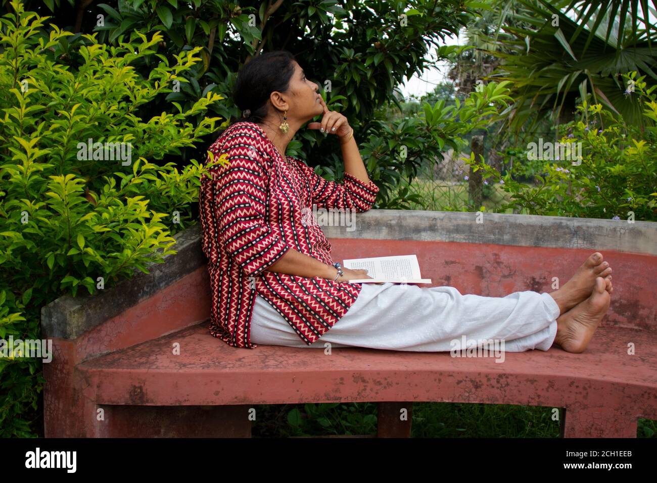 Une belle femme vêtue de vêtements décontractés, assise sur un banc rouge est en train de lire un livre et de penser à l'histoire dans un parc Banque D'Images