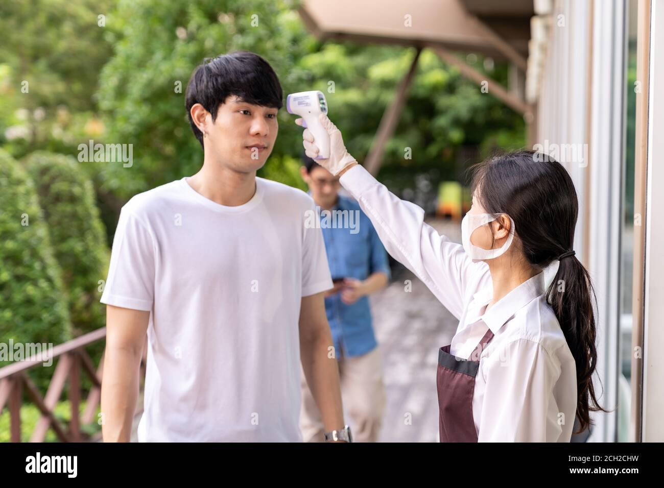 Serveuse asiatique avec masque facial prenant la température au client avant d'entrer dans le restaurant avec la file d'attente de distance sociale dans la file. Nouveau restaurant classique Banque D'Images