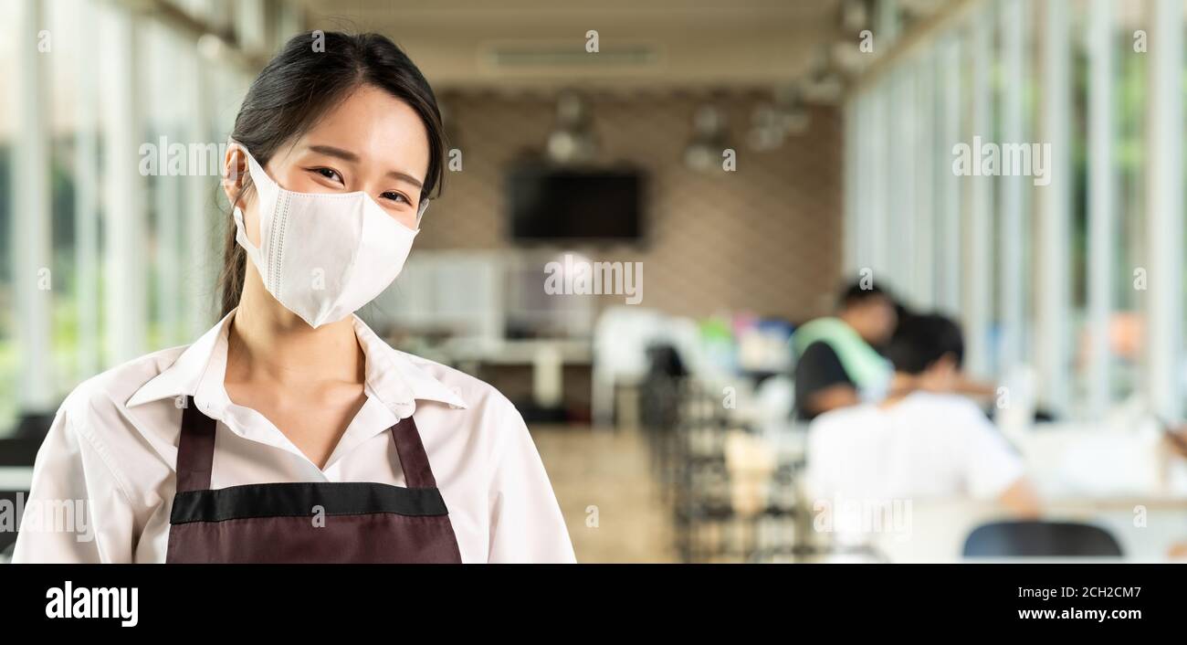 Portrait panoramique joli visage asiatique de serveuse avec le fond des clients assis dans la table de social distance dans le restaurant à l'intérieur. Nouveau repos normal Banque D'Images