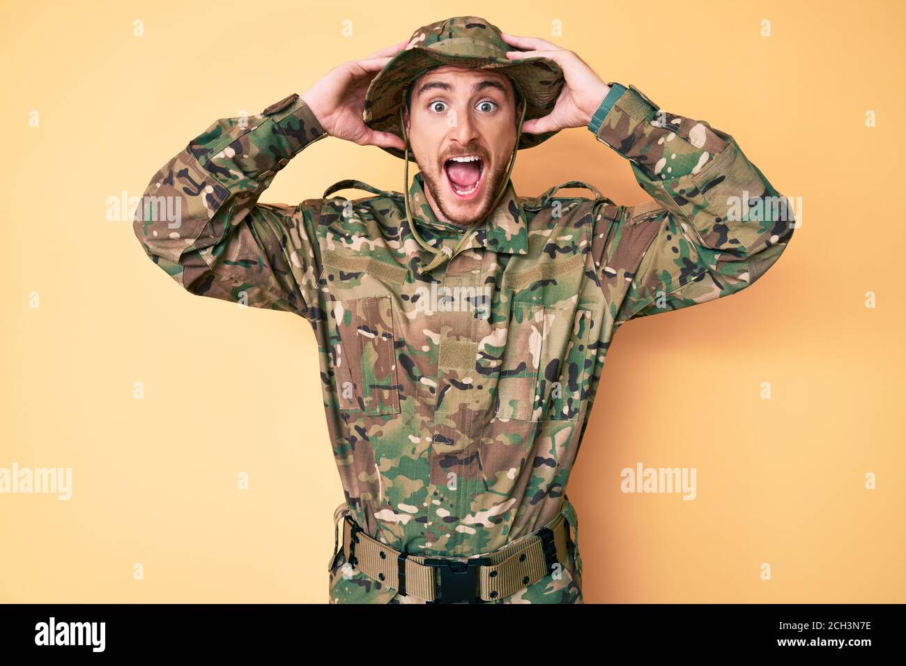 Jeune homme caucasien portant l'uniforme militaire de camouflage fou et effrayé avec les mains sur la tête, peur et surpris de choc avec la bouche ouverte Banque D'Images