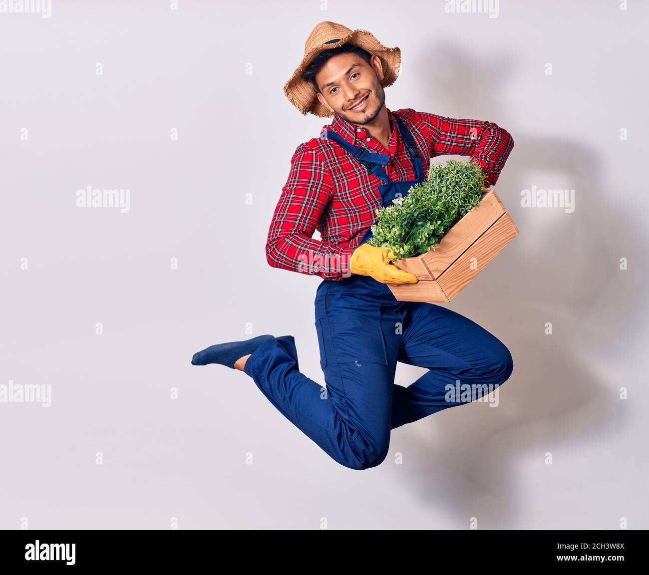 Jeune latins beau portant l'uniforme de fermier et chapeau souriant heureux. Sautant avec le sourire sur le visage plante pot en bois sur fond blanc isolé Banque D'Images