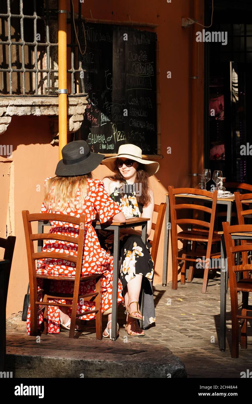 Deux jeunes femmes élégantes et attrayantes, portant des chapeaux et des lunettes de soleil à large bord, déjeunent à l'extérieur d'un restaurant typique dans le célèbre quartier touristique Trastevere Banque D'Images