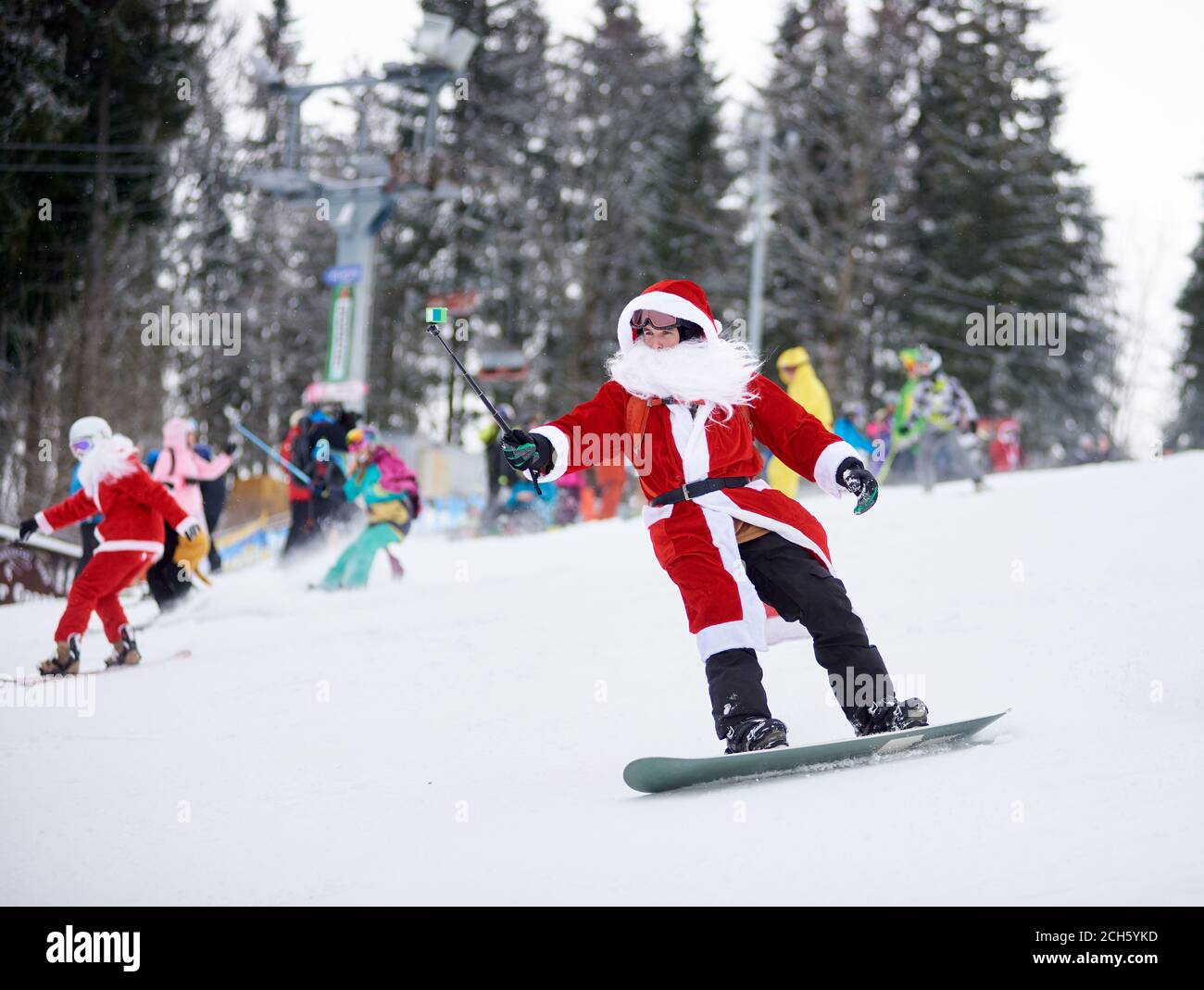 Bukovel, Ukraine - 09 décembre 2018 : homme snowboarder en costume de Père Noël avec barbe blanche tenant le monopied de selfie à la main, freeride sur le snowboard lors d'une compétition de freeride sur une piste de ski enneigée Banque D'Images