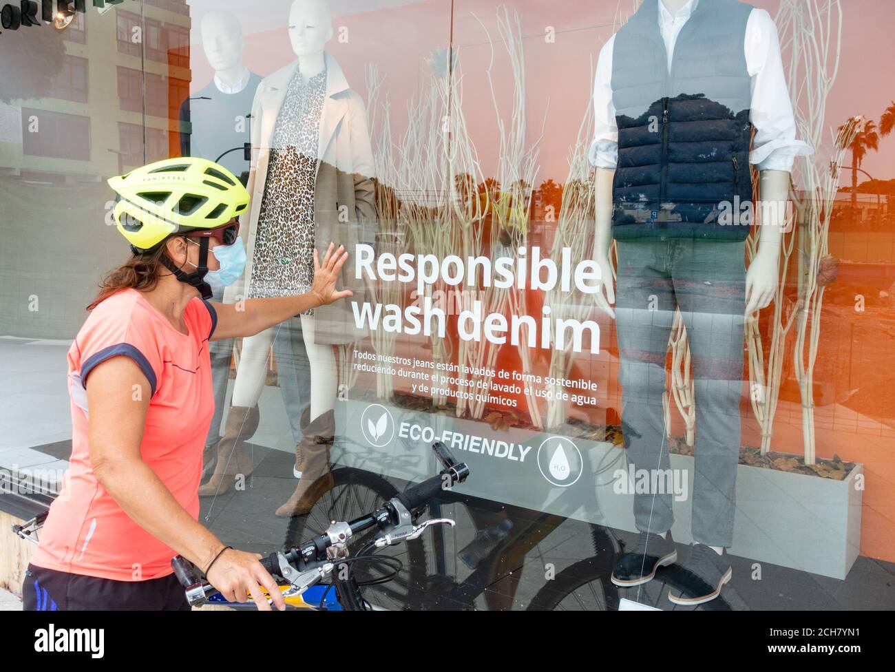 Femme cycliste portant un masque facial regardant dans la fenêtre du magasin avec un autocollant de denim de lavage responsable sur la fenêtre. Magasins de fenêtres, vêtements écologiques Banque D'Images