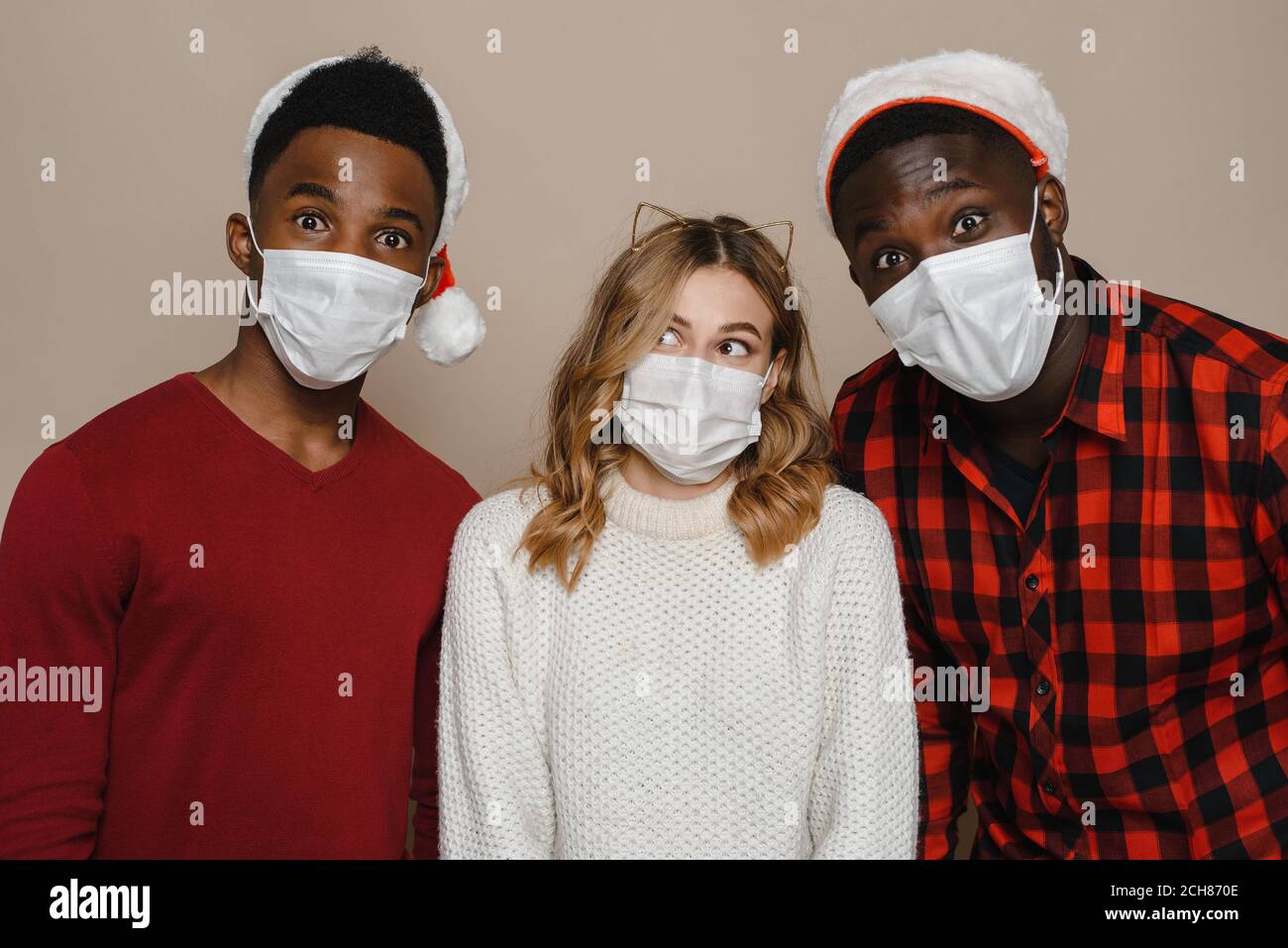 Trois jeunes amis portant des chapeaux du Père Noël et des masques médicaux. Restez à la maison et protégez-vous du coronavirus. Banque D'Images