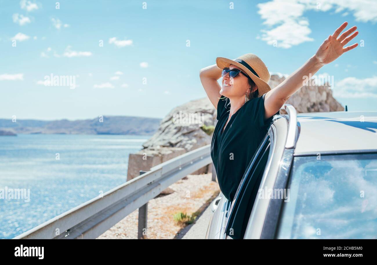 Portrait de femme joyeuse appréciant le voyage en bord de mer. Vêtue d'une robe noire, chapeau de paille, et lunettes de soleil elle a ouvert les bras larges Banque D'Images